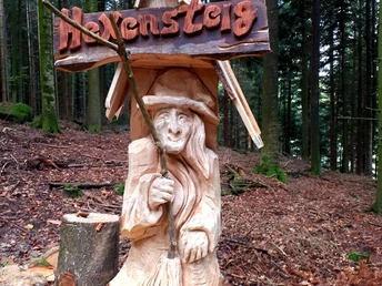 Die in einen Baumstamm geschnitzte Figur einer Hexe im Wald. Darüber ein Schild "Hexensteig"