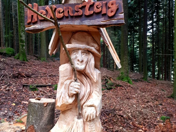 Die in einen Baumstamm geschnitzte Figur einer Hexe im Wald. Darüber ein Schild "Hexensteig"