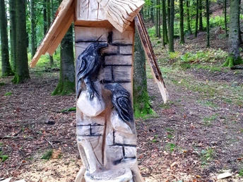 In einen Baumstamm geschnitzte Raben über einem Kessel, darüber ein Besen