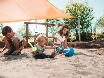 Gemeinsam spielen drei Kinder im Sankasten unter einem Sonnensegel