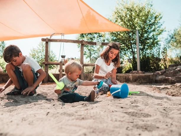 Gemeinsam spielen drei Kinder im Sankasten unter einem Sonnensegel