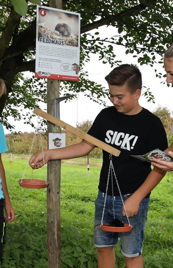 Zwei Jungs spielen an einer Station von Willi`s Wildtierpfad , eine Frau steht lachend dabei.