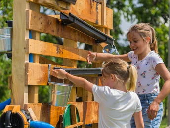 Zwei Mädchen spielen an einer Palette befestigtem Wasserspiel und lachen sich zu.