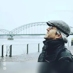 Ein älterer Mann mit grauem Bart und Brille blickt lächelnd am Fluss auf eine große Brücke.