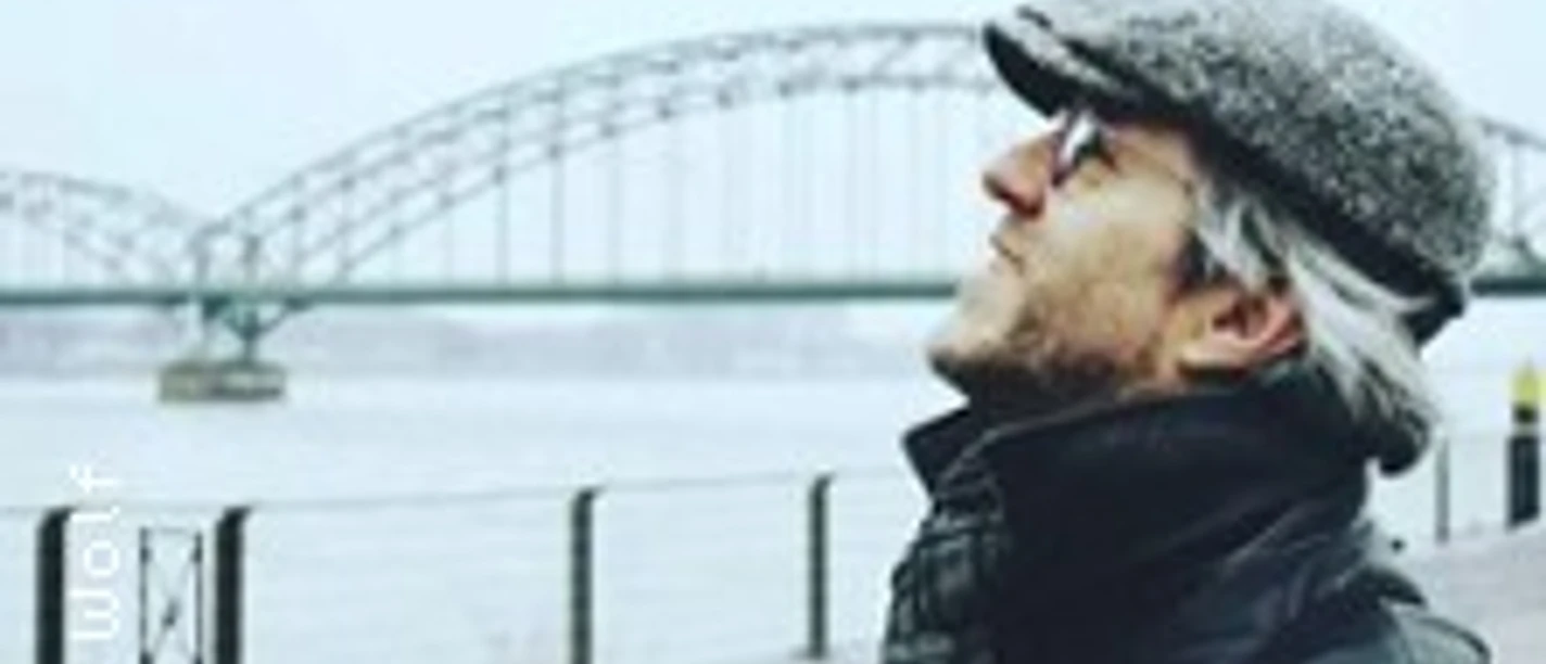 Ein älterer Mann mit grauem Bart und Brille blickt lächelnd am Fluss auf eine große Brücke.