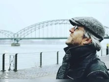 Ein älterer Mann mit grauem Bart und Brille blickt lächelnd am Fluss auf eine große Brücke.