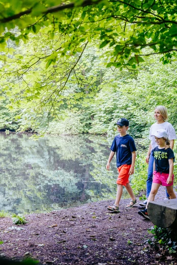 Familie spaziert im Grünen an einem ruhigen See entlang, umgeben von dichtem Baumbewuchs.
