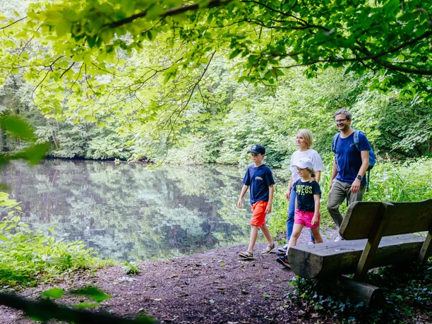 230626_besimmazhiqi-229.jpg Familie spaziert im Grünen an einem ruhigen See entlang, umgeben von dichtem Baumbewuchs.