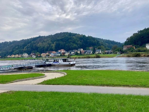 Fähre in Postelwitz Blick auf einen Fluss mit einem Boot und einer Uferpromenade, umgeben von grünen Wiesen und Hügeln.