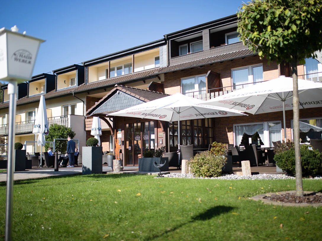 Ein dreistöckiges Hotel mit Außenbereich, Sonnenschirmen und Sitzgelegenheiten bei sonnigem Wetter.