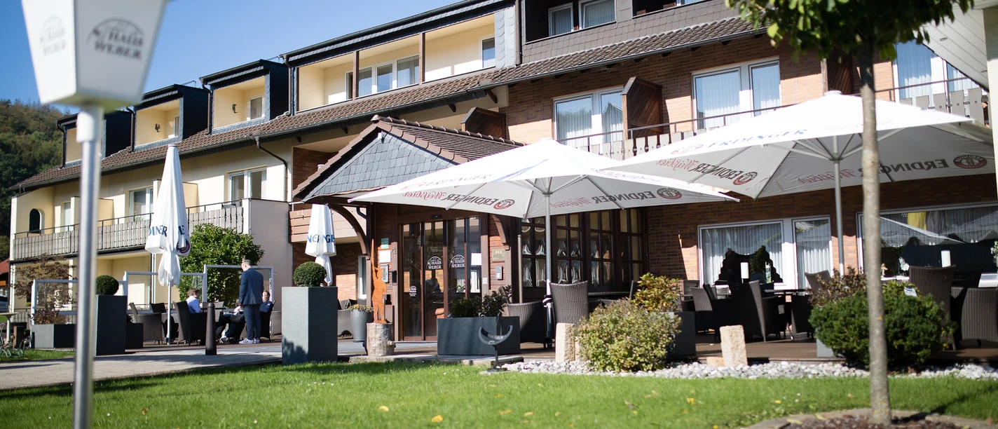 Ein dreistöckiges Hotel mit Außenbereich, Sonnenschirmen und Sitzgelegenheiten bei sonnigem Wetter.