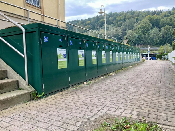 Fietsdozen_Bad_Schandau Groene fietsenstallingen met informatieposters staan langs een geplaveid pad, omringd door bomen en een bus op de achtergrond.