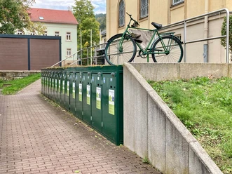 Fahrradboxen Bad Schandau Grünes Fahrrad lehnt an einem Geländer auf einer Betonmauer, daneben eine Reihe grüner Schließfächer entlang eines gepflasterten Weges.
