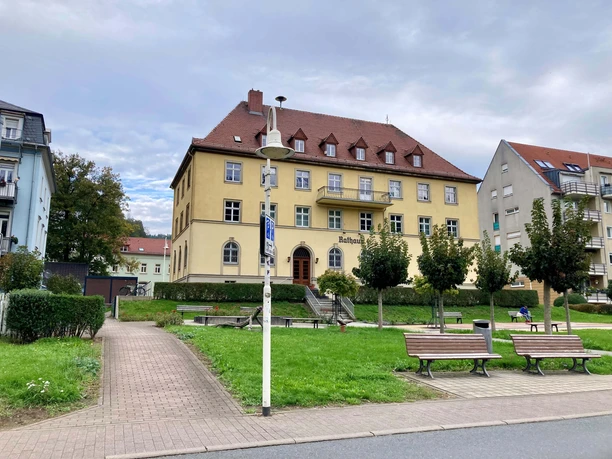 Přístup_k_garážím_pro_kola_z_Elbe Žlutá radnice s červenou střechou stojí mezi stromy a lavičkami v zeleném parku, obloha je zatažená.