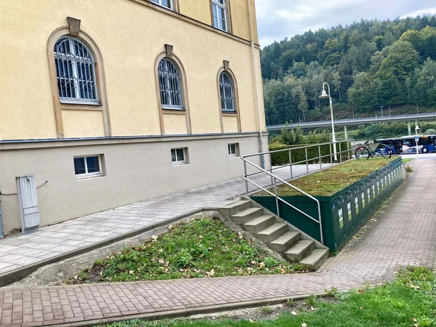 Yellow building with barred windows, next to it a bicycle on a ramp. Trees and a bus can be seen in the background.