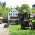 Historisches Mühlengebäude in grüner Waldlandschaft mit rustikalem Holzkarren und dekorativen Elementen.