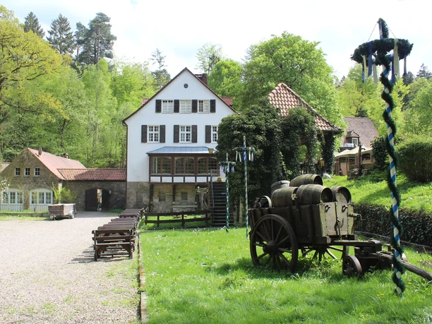 Historisches Mühlengebäude in grüner Waldlandschaft mit rustikalem Holzkarren und dekorativen Elementen.