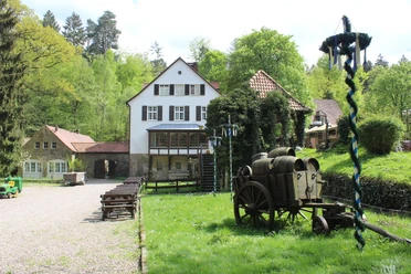 Silbermühle.JPG Historisches Mühlengebäude in grüner Waldlandschaft mit rustikalem Holzkarren und dekorativen Elementen.