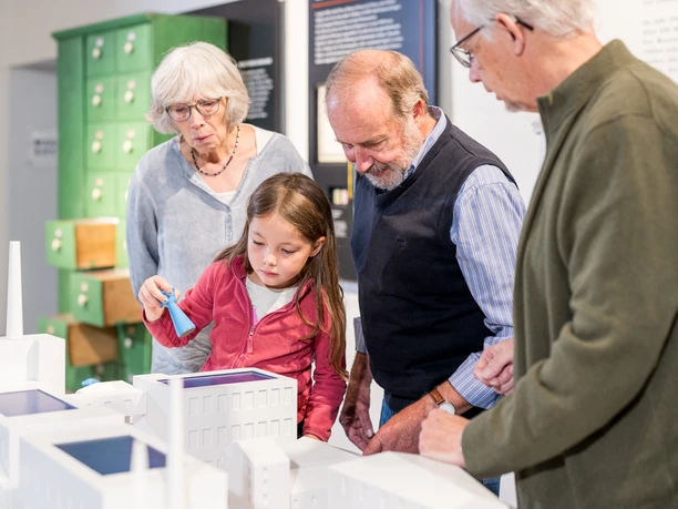 Am Fabrikmodell Eine Gruppe älterer Menschen und ein Kind betrachten gemeinsam ein architektonisches Modell in einem Museum.