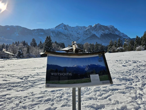 Blick auf die Alpspitze Schneebedecktes Feld mit Infotafel vor der Alpspitze unter klarem Winterhimmel