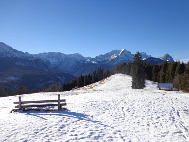 Eckbauer Verschneite Hügellandschaft mit Bänken vor bewaldeten Hängen und markanten Berggipfeln.