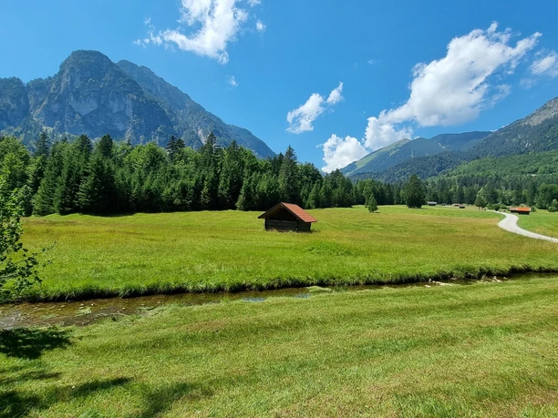 Blick auf die Reschbergwiesen Weite Wiese mit kleinem Bach, einzelner Holzhütte und hohen Bergen unter blauem Himmel