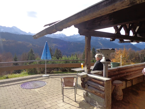 Pfeifferalm Terrasse mit Holzbank, Blick auf Berge und Wälder, Person sitzt mit Getränk unter Überdachung.