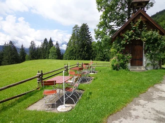 Biergarten Gschwandtnerbauer Holzbänke auf Wiese neben kleiner Kapelle vor Bergkulisse unter leicht bewölktem Himmel