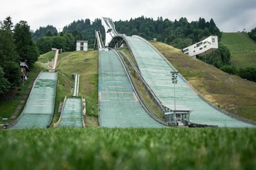 Olympia Skistadion Du siehst große Ski-Hänge für Sprung vor Wald und eine Hütte.
