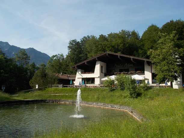 Aulealm, 5 Minuten vom Bobmuseum entfernt Du siehst eine Alm mit Teich und kleiner Fontäne vor Wald und Bergen.