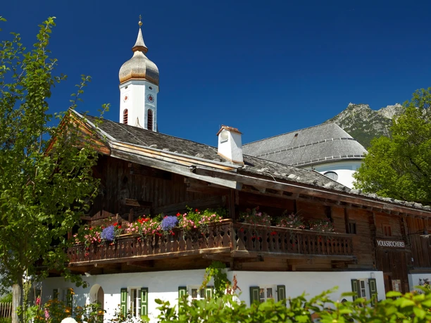 Polznkasparhaus mit der Kirche St. Martin Du siehst ein altes Holzhaus mit Blumen und eine Kirche dahinter.