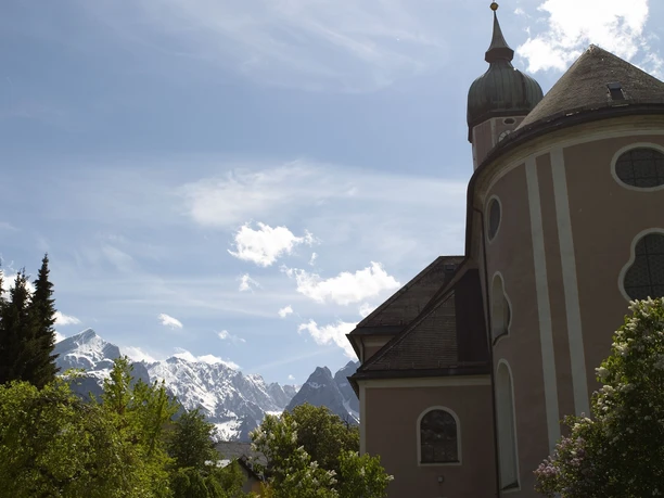 Pfarrkirche St. Martin im Garmischer Zentrum Du siehst eine Kirche mit Turm. Hinter ihr stehen hohe Berge.