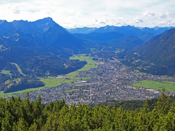 Panoramabild von Garmisch-Partenkirchen Du siehst Garmisch vor hohen Bergen. Grüne Wiesen umgeben die Stadt.