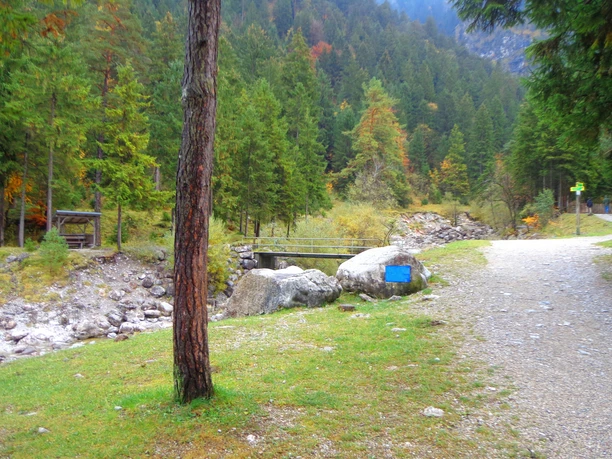 Farchant-Waldlehrpfad Breiter Waldweg mit Holzbrücke über steinigen Bach und einer überdachten Rastbank am Rand