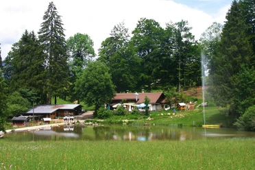 Kochelberg-Alm Almhütten am kleinen Weiher vor dichtem Wald mit hohem Wasserstrahl und Wiesen