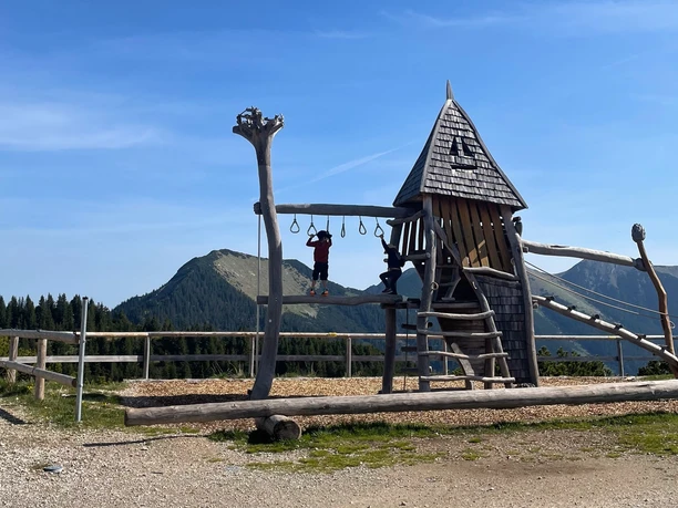 Wank Spielplatz Holzspielplatz am Berg mit zwei spielenden Kindern vor bewaldeten Gipfeln unter blauem Himmel