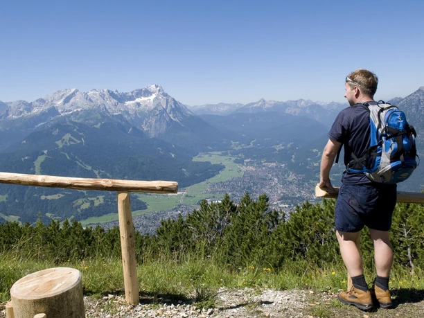 Wanderer blickt von einem Aussichtspunkt über ein weites Alpenpanorama mit Tälern und Gipfeln.