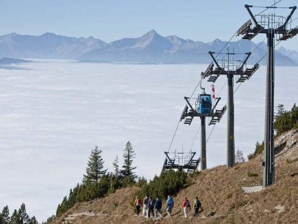 Wandernde auf einem Berghang neben einer Seilbahn über einem Wolkenmeer und fernen Gipfeln.