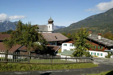 Blick auf Wamberg Alpendorf mit Kirchturm, traditionellen Häusern und umliegenden Bergen unter blauem Himmel.