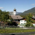 Blick auf Wamberg Alpendorf mit Kirchturm, traditionellen Häusern und umliegenden Bergen unter blauem Himmel.