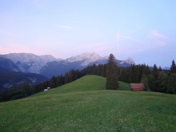 Eckbauer Weite Bergwiese mit zwei Holzhütten vor bewaldeten Hängen und hellen Gipfeln im Abendlicht