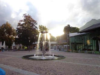 Richard-Strauss-Platz in Garmisch-Partenkirchen Du siehst einen Platz mit Brunnen. Die Sonne steht hinter Gebäuden.