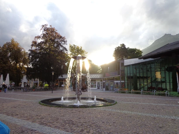 Richard-Strauss-Platz in Garmisch-Partenkirchen Du siehst einen Platz mit Brunnen. Die Sonne steht hinter Gebäuden.
