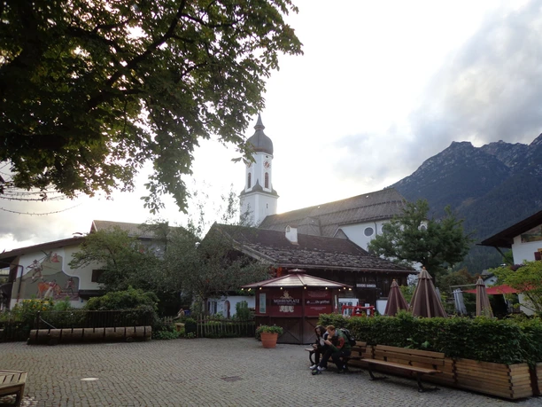 Polzenkasperhaus und Kirche St. Martin Du siehst ein Dorf mit Kirche, Haus, Bänken und Bergen im Hintergrund.