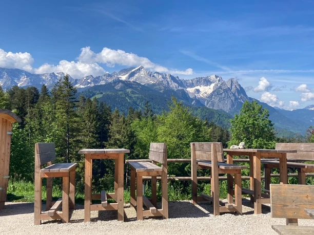 Ausblick von der Tannenhütte Holzbänke und Tische vor bewaldeten Hängen mit schneebedeckten Bergen unter blauem Himmel.