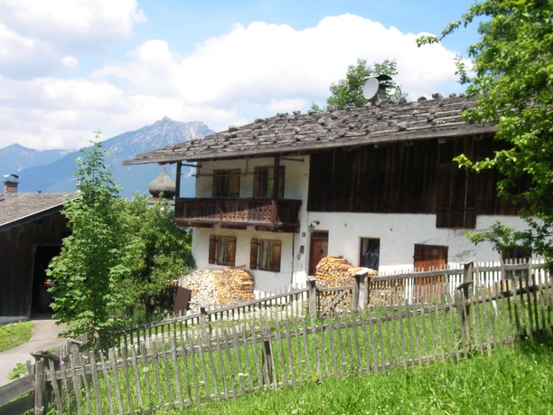 Bauernhaus in Wamberg Altes Bauernhaus mit Holzstapeln auf Hangwiese vor bewaldeten Bergen unter blauem Himmel.