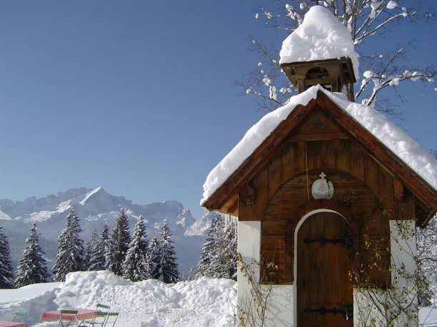 Kleine verschneite Kapelle Kleine verschneite Holzkapelle vor winterlichen Tannen und Bergkulisse unter blauem Himmel