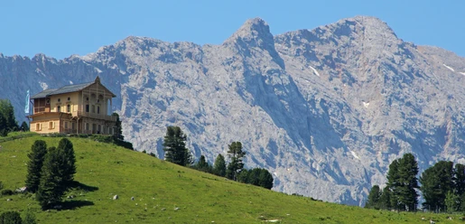 Holzhaus auf grünem Hang vor steiler Felswand unter klarem Himmel