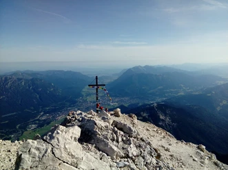 Alpspitze Du siehst helle Felsen mit einem Gipfelkreuz und weite Berge im klaren Licht.