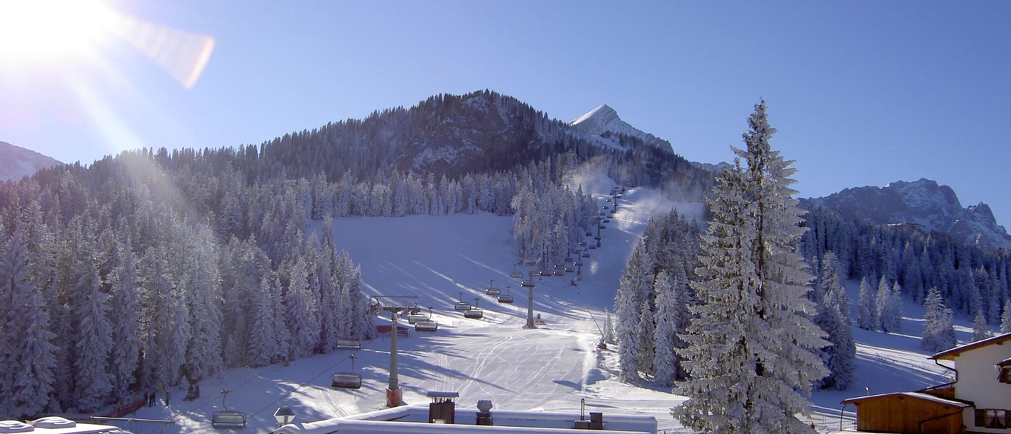 Kreuzwankl-Ski-Express Du siehst eine sonnige Skipiste mit Liften und schneebedeckten Bäumen.